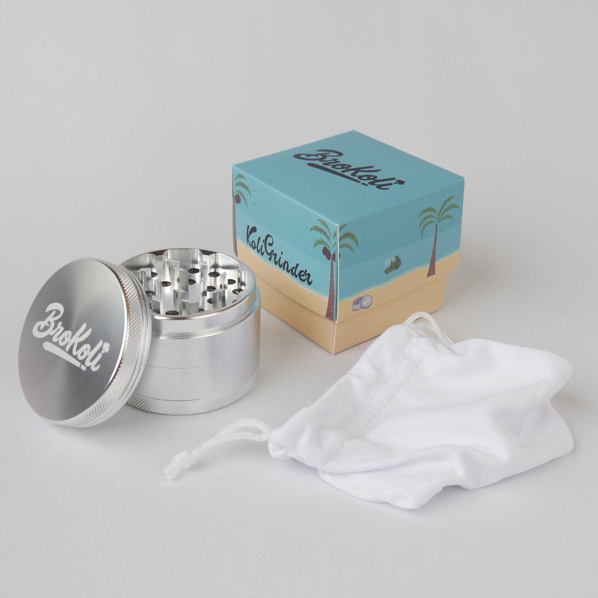 Der KoliGrinder von BroKoli aus Aluminium mit geöffneter Mahlkammer und graviertem Logo auf dem Deckel. Daneben liegt die passende Verpackung mit tropischem Design, Palmen und Strandmotiv sowie ein weißer Stoffbeutel zur sicheren Aufbewahrung.