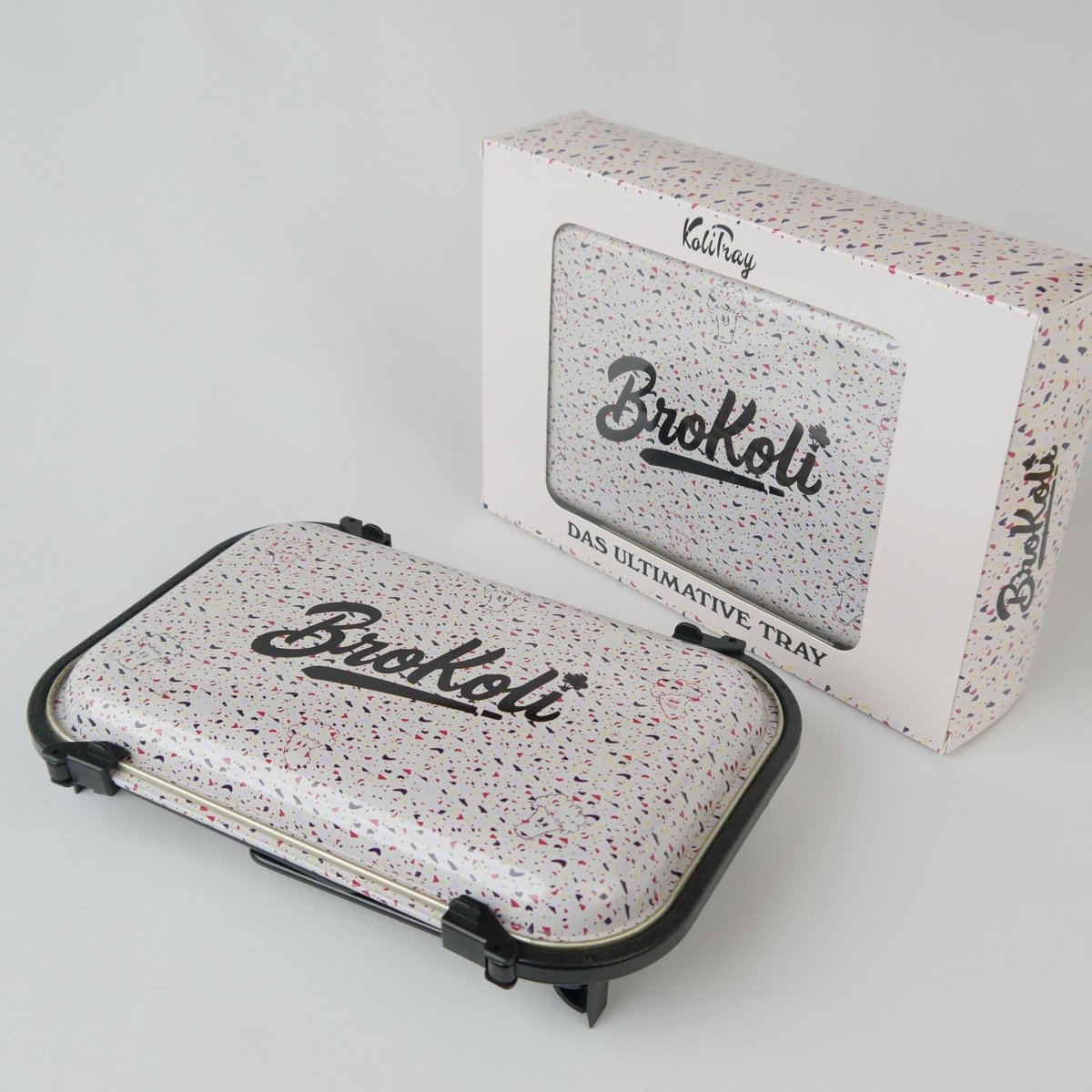 Das KoliTray von BroKoli mit Verpackung. Das faltbare und verschließbare Tray hat ein Terrazzo-Design mit bunten Sprenkeln und das BroKoli-Logo auf dem Deckel. Die Verpackung zeigt das gleiche Design mit Sichtfenster und der Aufschrift „Das ultimative Tray“.